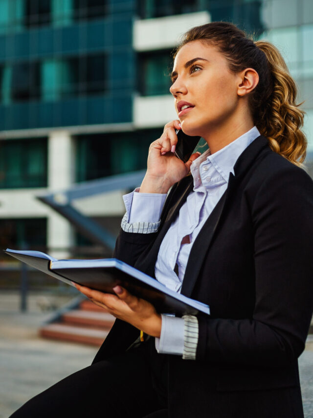 Successful businesswoman or entrepreneur taking notes and talking on cellphone outdoors.